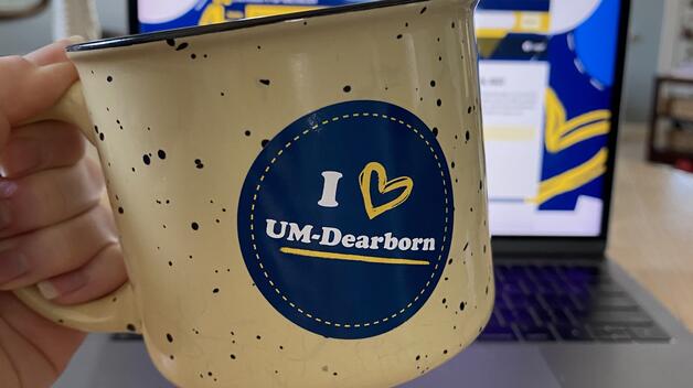 A hand holds a mug with a "I heart UM-Dearborn" sticker on it.